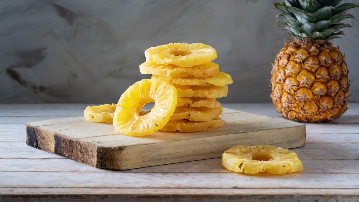 Getrocknete Ananasringe auf einem Holzbrett, frische Ananas im Hintergrund.